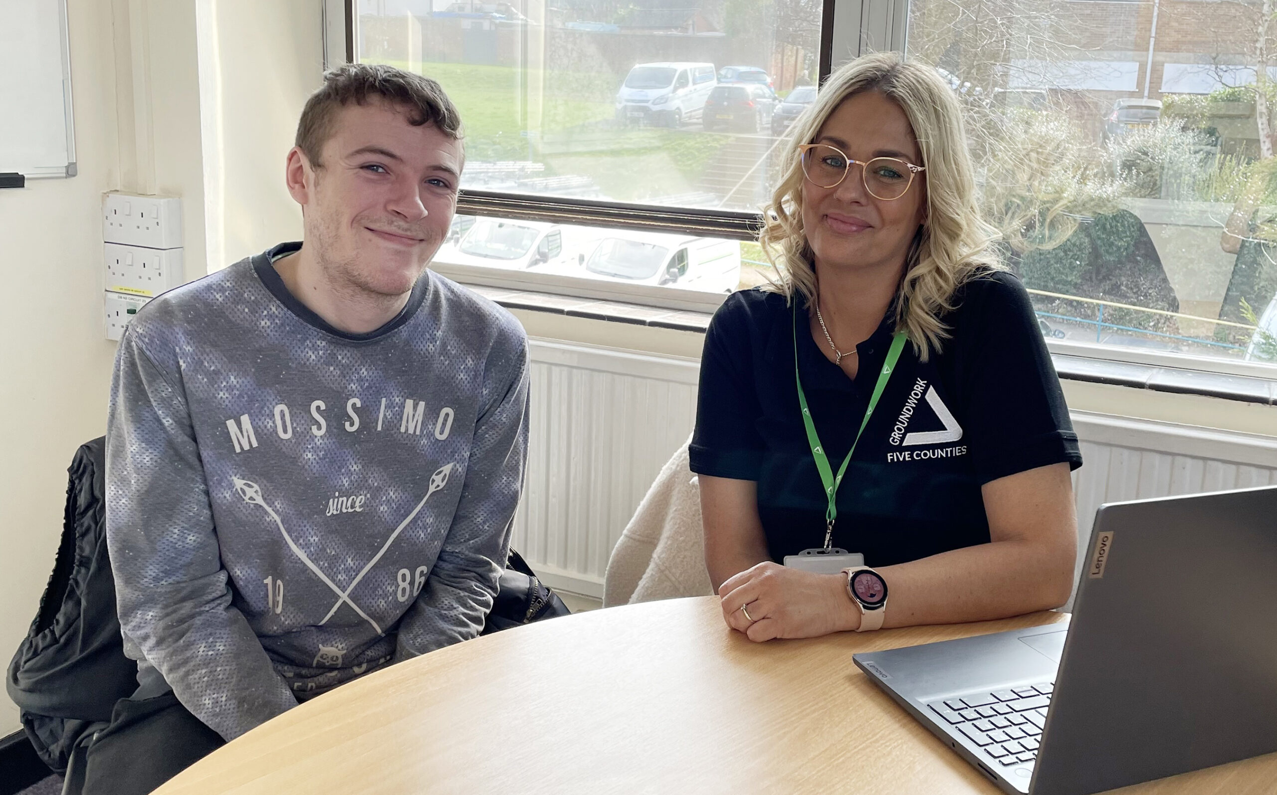 Young man wearing grey top and blonde woman in black top with green lanyard sitting at desk with laptop, smiling at camera.