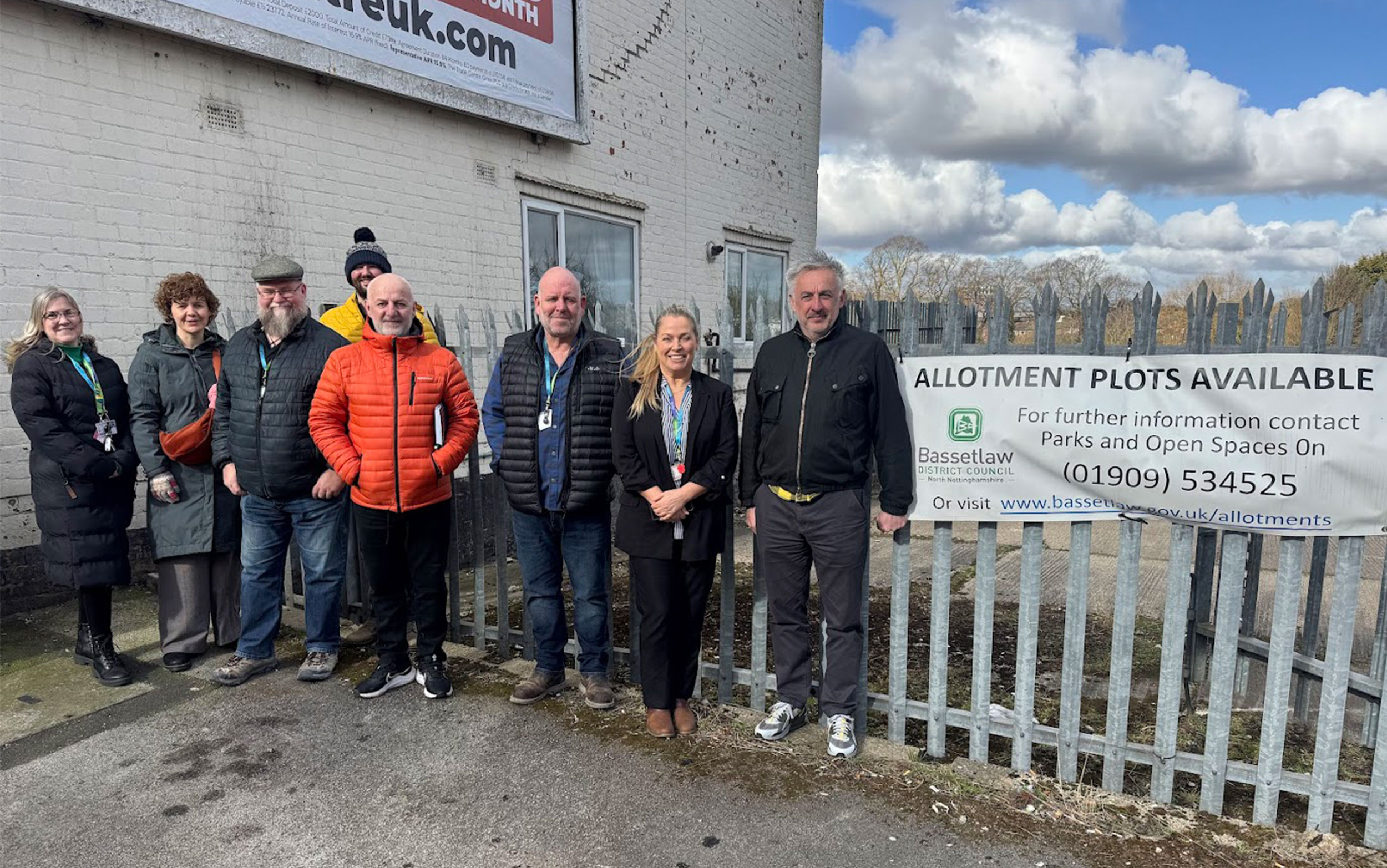 Group of people standing by metal gates with white banner with black text "Allotment Plots available".