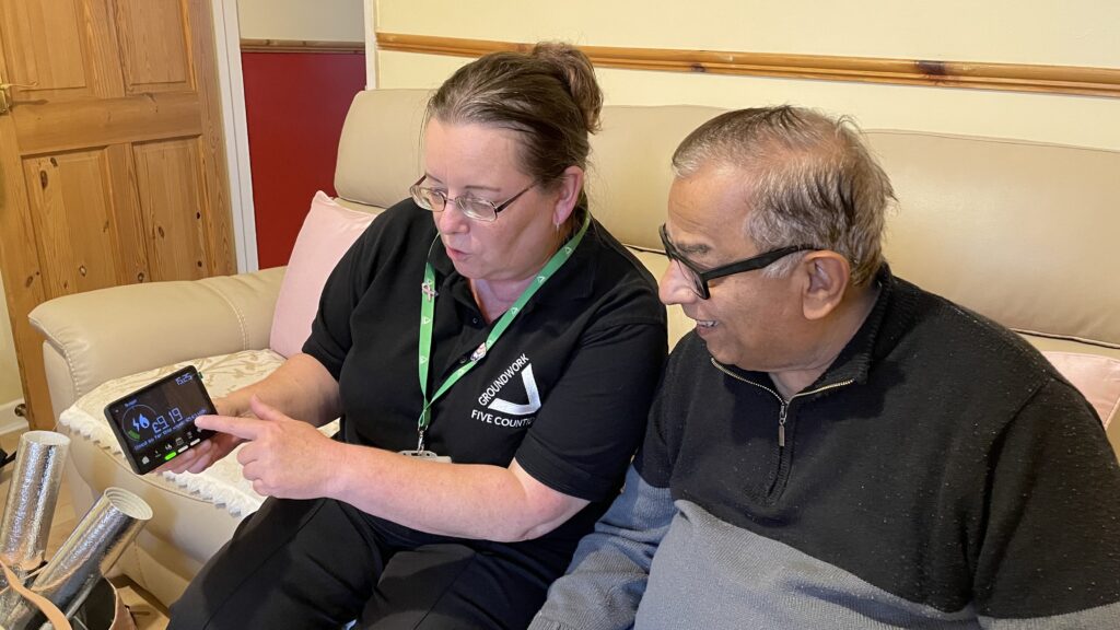Woman wearing black top with white Groundwork logo, showing man a smart meter. They're sitting on a beige sofa in a room.