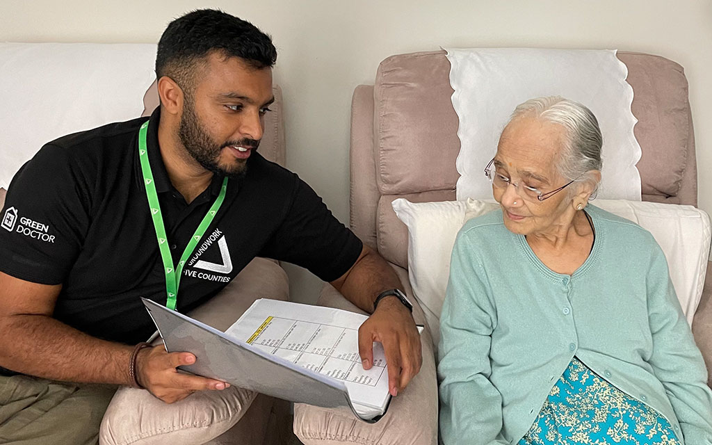 Young man wearing black top with white Green Doctor and Groundwork logo showing paper to elderly woman in blue. Both are sitting in chair