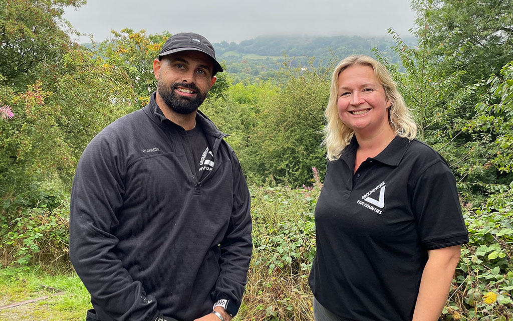 Man and woman wearing black tops with white Groundwork Five Counties logos, with greenery in background.