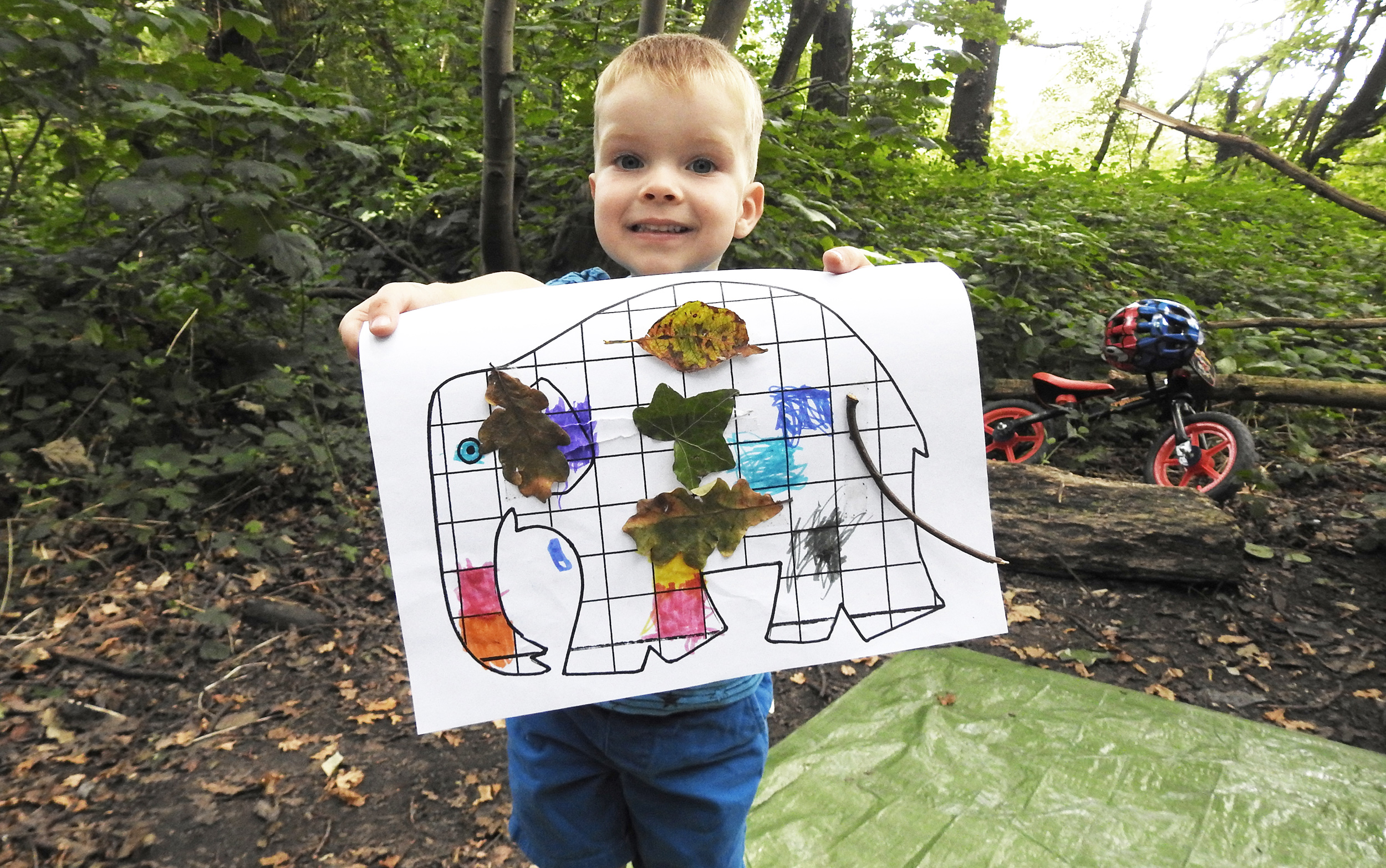 Young boy holding paper with elephant drawing and leaves attached, smiling at camera. Outdoor setting with bicycle in background.