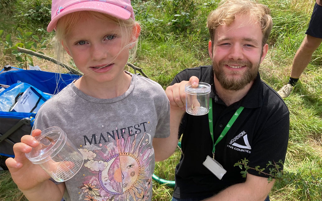 Young girl and man holding clear pots with insects, in outdoor setting.