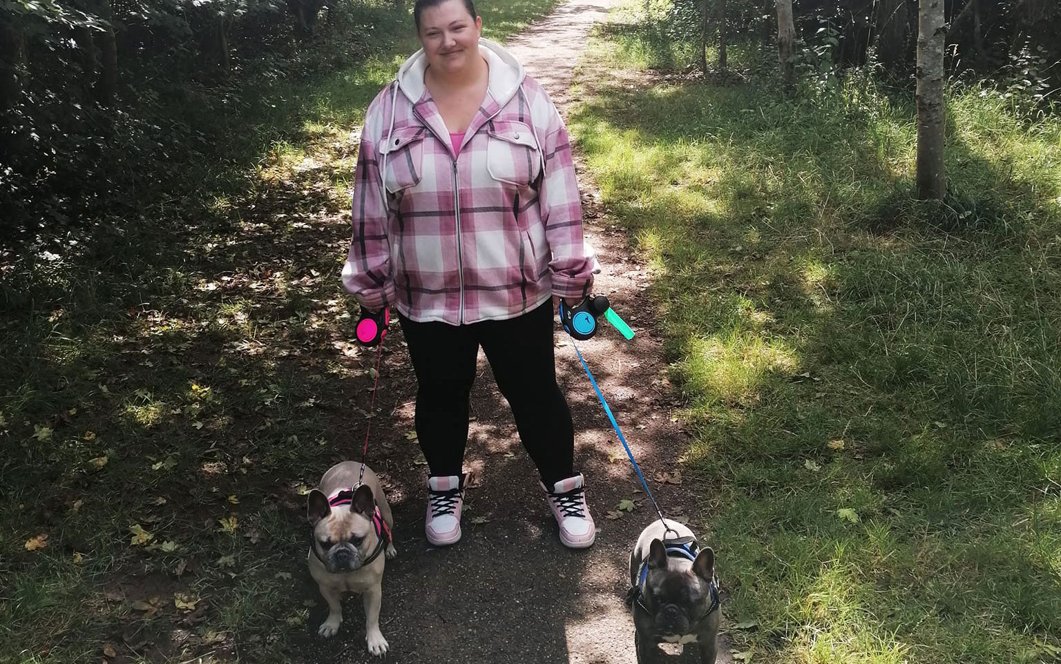Dark haired woman wearing pink coat with two dogs on leashes in nature, smiling at camera.