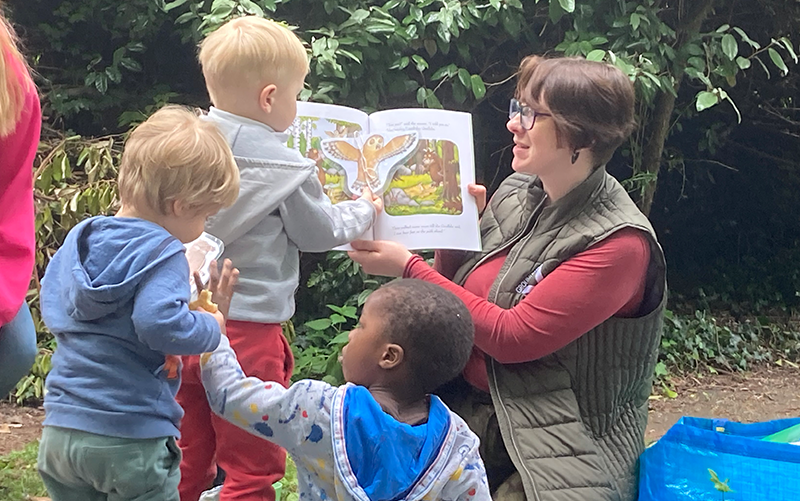 Woman reading from picture book with children around, in outdoor setting.